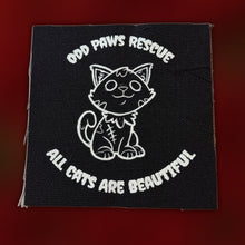 Load image into Gallery viewer, A square black fabric patch with white embroidered text reading 'ODD PAWS RESCUE' at the top and 'ALL CATS ARE BEAUTIFUL' at the bottom, surrounding a white line-art cat illustration. The patch is photographed against a dark red gradient background.
